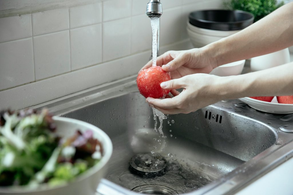 Hands washing a fresh red apple under running water at a kitchen sink.